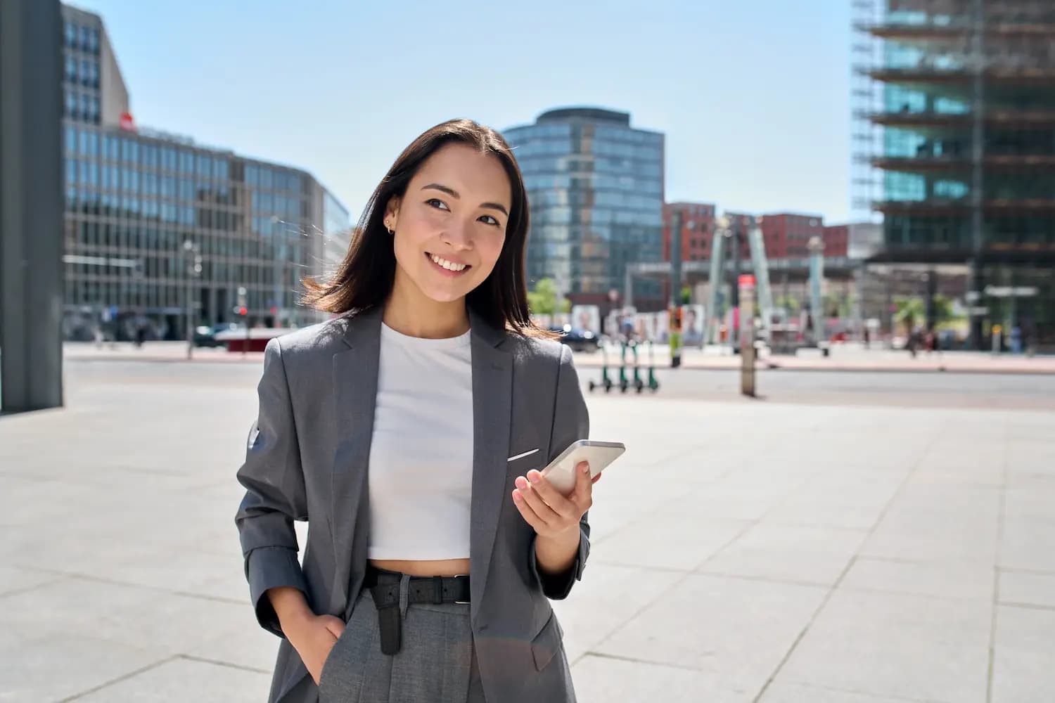 Young smiling elegant Asian busy business woman leader wearing suit standing in big city using cell phone platform applications. Smiling woman holding smartphone walking on street outdoors.